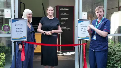 Sue Johnson (68) who is wearing a longdark blue summer crinkly dress, with a black jacket. She has white hair in a Cleopatra bob. She is smiling. Her daughter, Rachael is next to her. She is wearing a black dress, with dark hair tied back. She is smiling and about to cut a red ribbon to open the unit. Next to her is a consultant nurse who works at the unit. She is wearing dark blue hospital scrubs and has short light brown hair.