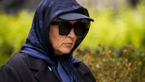 PA Media A woman wearing large sunglasses, a sun hat and a scarf is walking down the street.