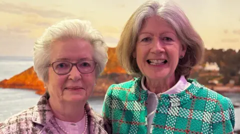 Pamela has short white hair and is wearing glasses and a cardigan. Martina has shoulder-length brown hair and is wearing a chequered jacket.