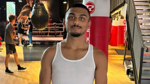 Roshan standing in front of the boxing gym, wearing a white vest, black shorts and white boxing gloves with orange thumbs.