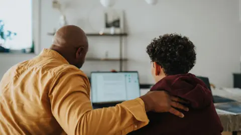A stock image of a man and a boy using a computer, taken from behind. The man has his arm around the boy. It is unclear what is on the screen.