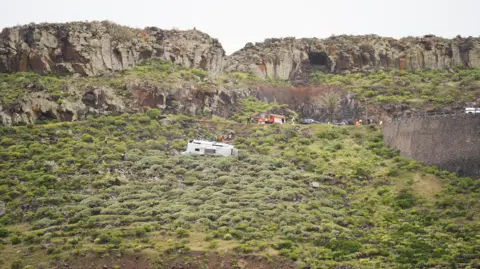 Europa Press Canarias via Getty Images A bus seen on its side among shrubbery next to a road, with rocks in the background