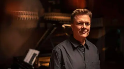 Daniel Lewis/Zurich Toby Spence, a man wearing a dark shirt, stands in front of a black grand piano looking into the camera