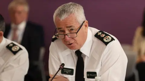 PA Media Jon Boutcher leans into a microphone while speaking at the Policing Board. he has white hair and is wearing black-rimmed glasses and is wearing a white shirt with PSNI badges on it