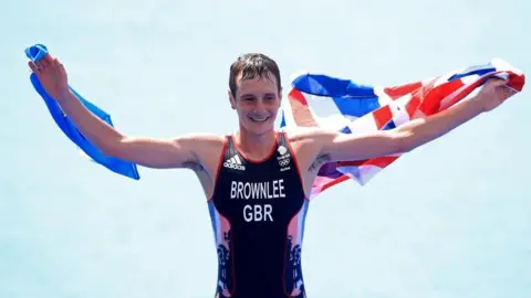 Alistair Brownlee holds two flags: the Union Jack and Yorkshire's white rose flag after winning gold in the Rio OIympics. He is a man with short dark hair wearing a black and red vest. 