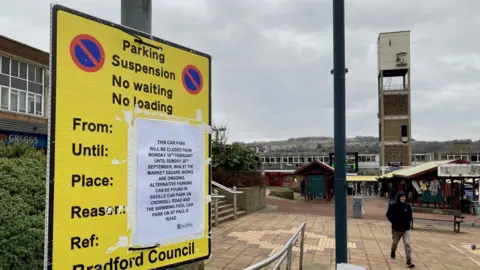 A yellow and black parking restrictions sign with piece of paper taped to it outlining how long the works will last.  