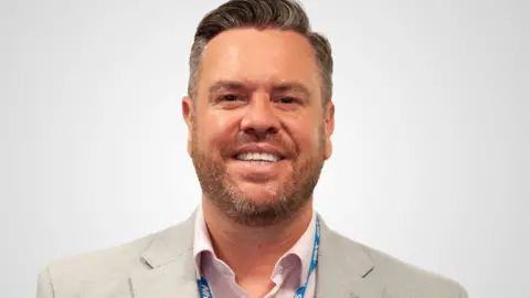 NHS Lancashire and South Cumbria ICB Craig Harris, chief commissioning officer for NHS Lancashire and South Cumbria ICB with grey hair and beard, wearing a beige blazer and pale lilac shirt with a blue NHS lanyard. He is smiling.
