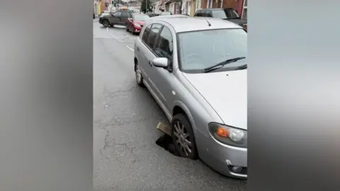 A silver car has its front driver's side wheel stuck in a sinkhole, which has appeared on a residential road. There is queueing traffic behind the car, and an SUV-type car can be seen turning around.