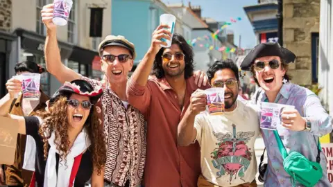 Tom Warner A group of people stands close together in a narrow street decorated with colourful bunting, holding up reusable festival cups towards the camera. Shop signs and pastel-coloured buildings line the street behind them, adding to the bright and busy festival atmosphere.
