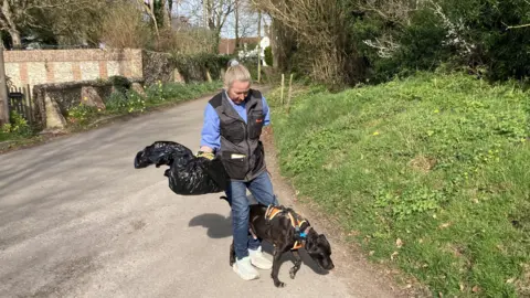 Diane Kasperowicz and her black sniffer dog Louis walking on a path looking around. Diane is holding a black bin bag in her hands.