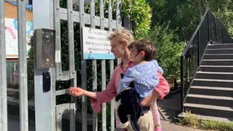 Luisa, a woman with short blonde hair and a pink top, holding her daughter Leila while opening a metal gate that accesses the nursery