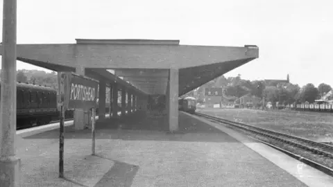 John Thorn Old archive picture in black and white of old railway station. There's a big sign saying Portishead indicating the stations names. There's a platform with several trains on the track with trees and a church in the distance.