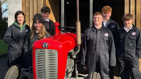 A red vintage tractor is surrounded by teenagers in boiler suits - three on each side, one girl on the left at the front has a black beanie on.