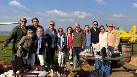 Mr Fox with all of his family, behind them is an airfield with vintage planes on.