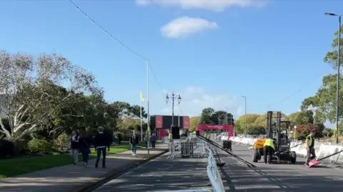 Workers dismantling the finish line of the Great South Run. People are walking on the nearby pavement. It's a sunny day.