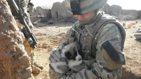 Family Handout Nadine Askew wearing a camouflage patterned military uniform and helmet, standing in a dry and dusty warzone surrounded by abandoned buildings. She is holding a small black and white puppy, which has curled up in her arms.