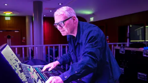 A man with silver hair and black glasses leans over an audio mixing desk in a dark room.
