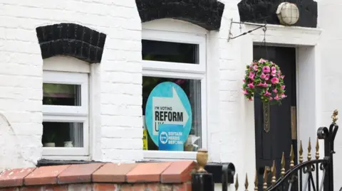 PA Media A white painted brick house with a hanging flower basket over its door and a Reform UK poster in the window.
