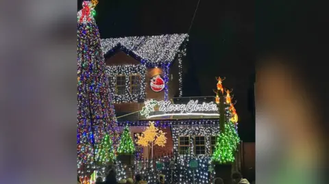 Paul Molyneux The same house is shown from a wider angle, featuring a huge illuminated tree and a glowing “Merry Christmas” sign. Reindeer figures appear on raised poles, and green light-up trees line the front. The entire display sparkles with white and colored lights.