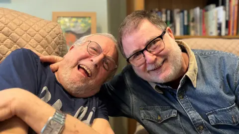 BBC Bernard and Danny Mendoza, both smiling on camera at a care house. Danny has placed his hand around his father.