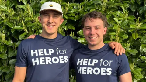 Two young men standing in front of some foliage, with their arms around each other. The one on the left is taller and wearing a white baseball cap, while the other has brown hair. Both are wearing dark-blue t-shirts with Help for Heroes written on them in white letters.
