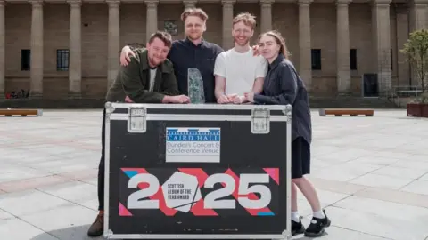Three young men and a woman stand outside a large hall in a city square. They are leaning on a large packing case with the words Scottish Album of the Year 2025.