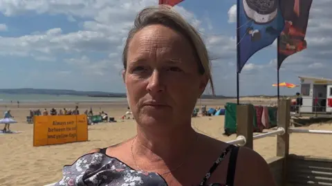 George Carden/BBC A woman with long, blonde hair looking past the camera. She is in front of a sandy beach.