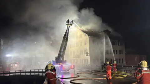 Graubünden cantonal police The building as seen from a bridge crossing a river. Three streams of water from firefighting crews douse the building. Smoke rises from the building - at night. 