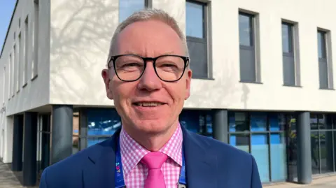 Andrew Morgan, who is looking into the camera and smiling. He is wearing a pink and white checked shirt, a pink tie, and a dark blue blazer. He has short, light blond hair and is wearing a pair of dark-framed glasses. Morgan is photographed outside a hospital building, which is a two-storey structure. The lower storey has dark grey pillars and floor-to-ceiling windows, whilst the upper floor is painted cream and has about eight, narrow, grey-framed windows visible. 