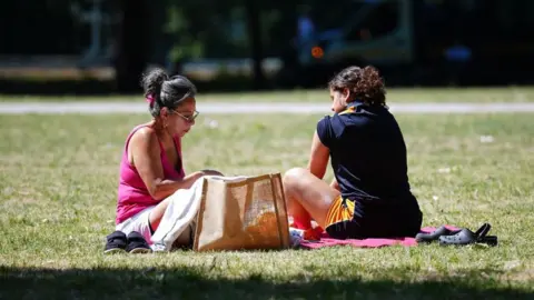 A woman with long hair pulled up in a bun, wearing a pink top, sits on the grass opposite another woman, wearing a dark top and shorts, in Vauxhall Park in London.