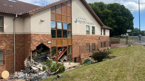 A large hole at the bottom left of a brick and white stone building next to a window. Bricks and debris from the wall are scattered all around and in the bushes and grass in front of the building. 