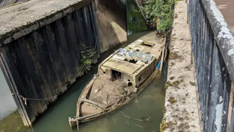 Bath and North East Somerset Council A partially submerged boat wreck covered in mud and appearing to block a lock in the river