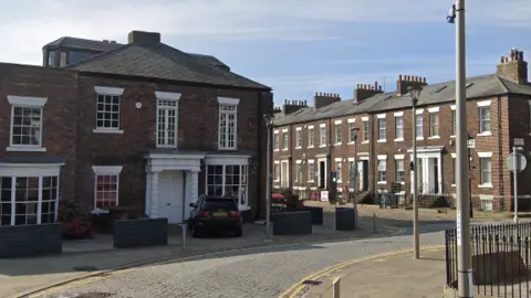 A street view of brick, terraced housing with white windows and doors. The road is cobbled.