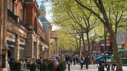Getty Images Bridge Street in Peterborough. There are people sitting outside cafes and others walking along the street. Part of the Town Hall can be seen. 