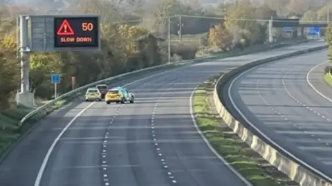 A closed stretch of the M5, completely clear apart from two police vehicles and another car, with a gantry sign above saying "50; Slow down"