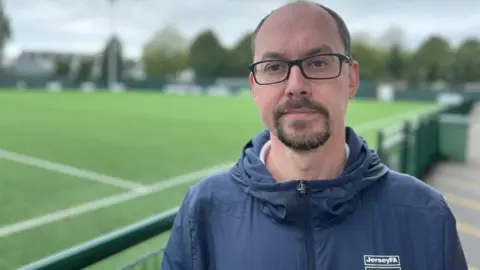 BBC Tim Pryor is wearing a blue waterproof top with the logo of Jersey FA on it. He is standing in front of the football pitch at Springfield Park.