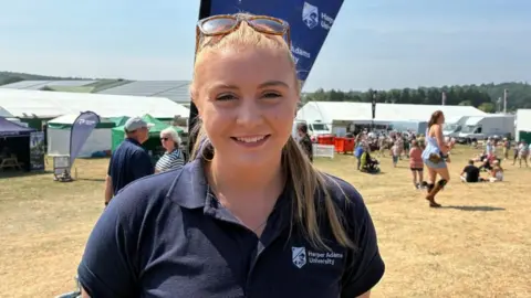 A young woman with blonde hair tied back in a ponytail smiles. She wears sunglasses on her head and a navy blue polo shirt with a white Harper Adams University logo on the chest.