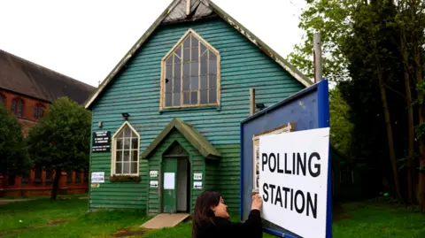 A wooden church painted blue and green and with pointed windows has a blue sign outside, a woman with shoulder length brown hair is attaching a white poster that says 'polling station' in black writing on it. It is a stock image from a previous election in Birmingham