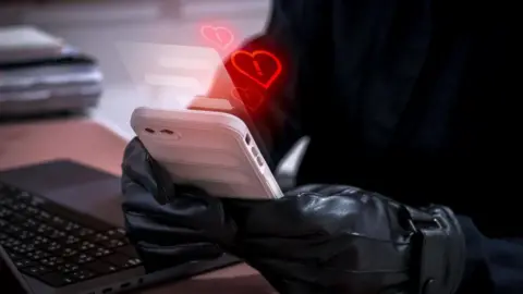 Romance scam concept showing a cybercriminal wearing black gloves using a white smartphone to deceive victims with fake love messages with red hearts with exclamation marks coming from the phone. A laptop is next to them on a desk.