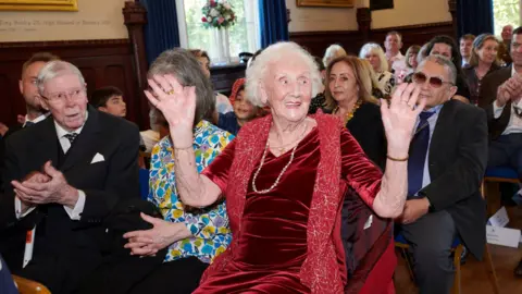 Banbury Town Council Ena Bowley wearing a red dress and holding her hands up and smiling at people. She is surrounded by crowds at an event in a hall.