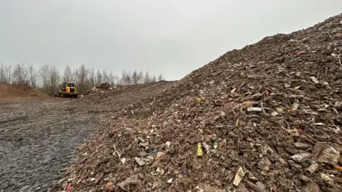 A close up image of a large pile of waste which extends out into the distance of the photograph. The waste is made up of wood, plastic and other bits of household waste. There is a yellow JCB truck in the background. 