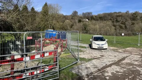 BBC/Phil Harrison A white car sits at the entrance to a field. There is heras fencing and red and white security fencing surrounding the field. 