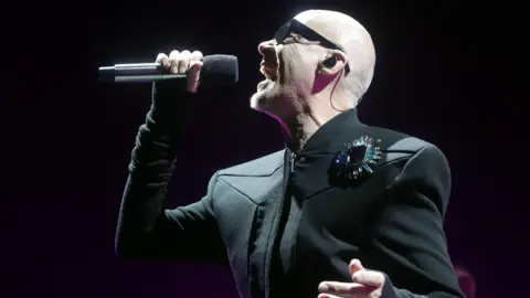 The Human League's Philip Oakey is wearing a black jacket, sunglasses and is singing into a microphone.