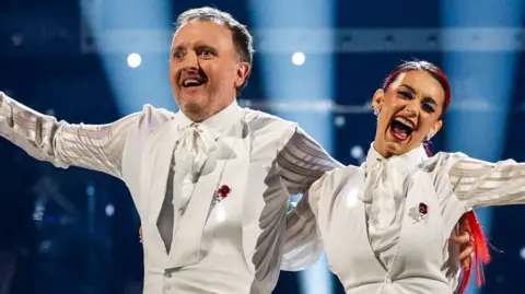 Chris McCausland and partner Dianne Buswell during the Strictly live show on 9 November. Wearing white suit and holding their arms out.