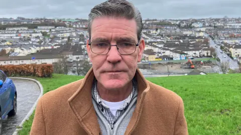 A man in a tan coloured coat is wearing a greay jumper and shirt, and a white t-shairt. he is standing on an area of grass , behind him is a cityscape. He is wearing eye glasses and has sort greying hair.