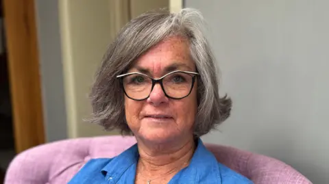 Sue Angus is looking at the camera. She has grey hair and glasses. She is wearing a blue blouse and is sitting on a pink chair. She is pictured from the shoulders up.
