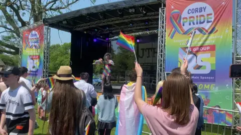 Patrycja Boryka/BBC A temporary black stage with Corby Pride billboard on either side. A person is performing at the front of the stage in a rainbow coloured outfit. There are people watching, some of whom are holding Pride flags.
