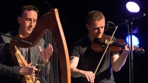 Getty Images Two men sitting side by side as they play their instruments. Mr Rooney on the left plays his brown wooden harp, he has short dark hair and is wearing a dark grey suit with a patterned tie. On the right is Mr Byrne with short brown hair and facial hair who is wearing a black t-shirt and is playing a fiddle. Behind then both is a black backdrop and two coloured stage lights.