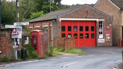 Cranborne fire station is a small brick building on a bend in a village road, with an adjacent red phone box and a signpost giving directions to Dorset towns and villages.