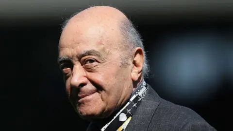 A close up on Mohamed Al Fayed, who is wearing a grey suit and has a bald head. The background is dark and blurred and his head it tilted so he is looking at the camera.